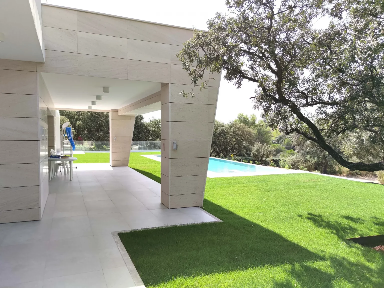 Zona de porche exterior techado con pilares de piedra, ideal para comedor de verano, con vistas hacia el césped.