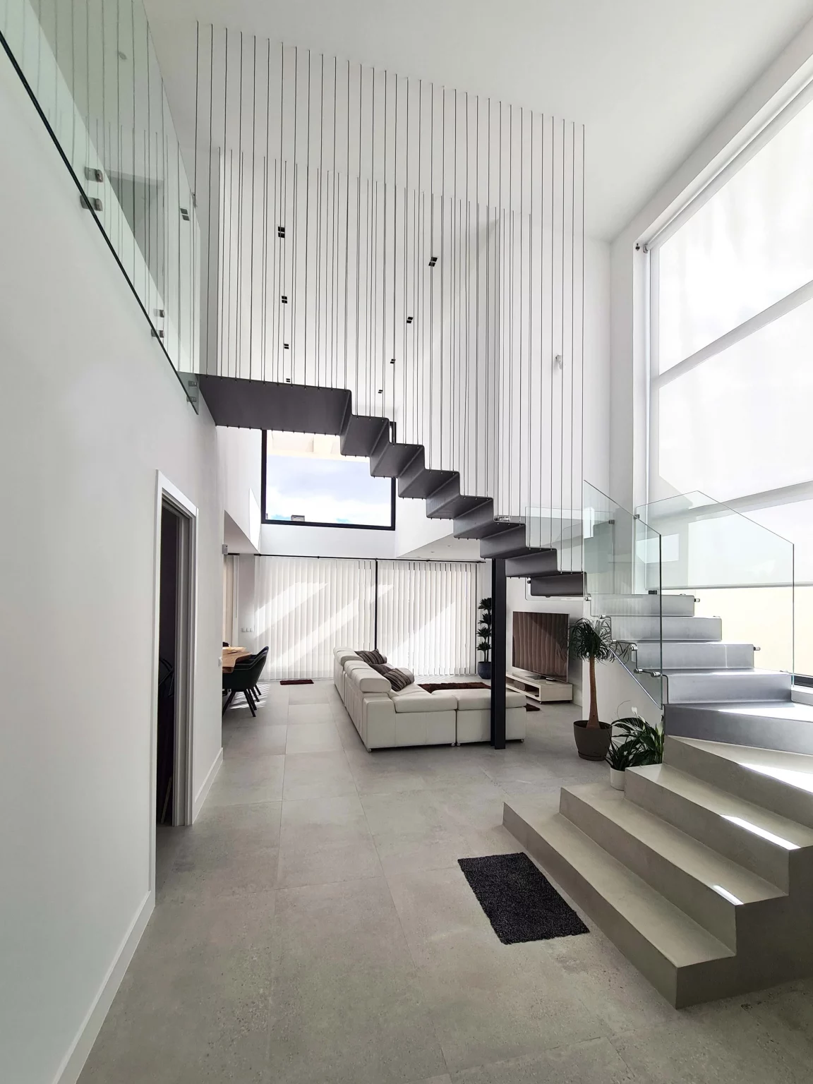 Interior de un salón luminoso de doble altura con escalera metálica flotante y barandilla de cristal en Villa V25.