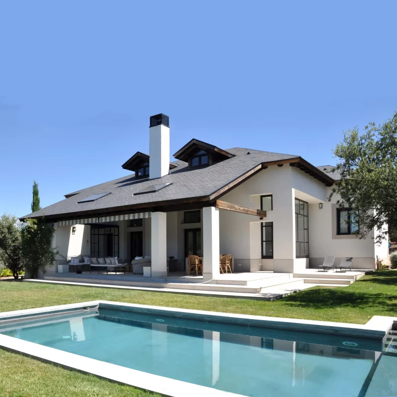 Vivienda de cubierta inclinada con buhardillas, porche con vigas de madera y piscina rectangular sobre el césped